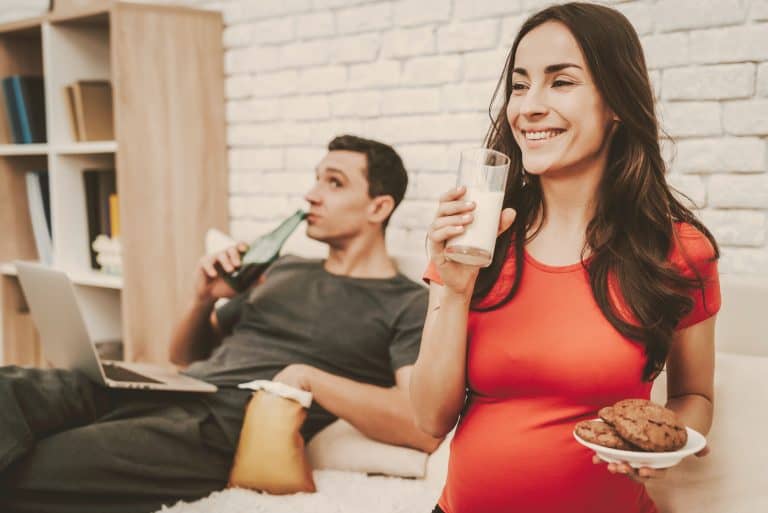 Couple Is Sitting On Couch. Woman Is Drinking A Milk And Eating A Cookie. Woman Is Pregnant And Smil