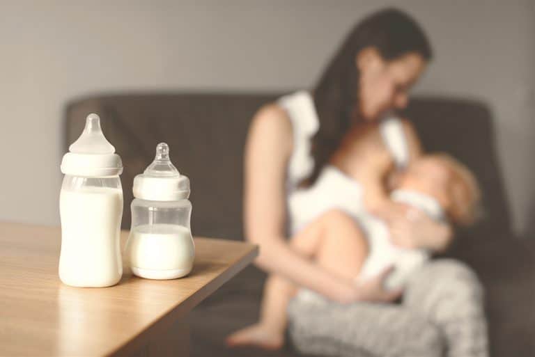 Bottles With Breast Milk On The Background Of Mother Holding In Her Hands And Breastfeeding Baby. Ma