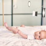 Adorable Baby Boy Infant In White Sunny Bedroom Lying And Looks At Munari Montessori Mobile.