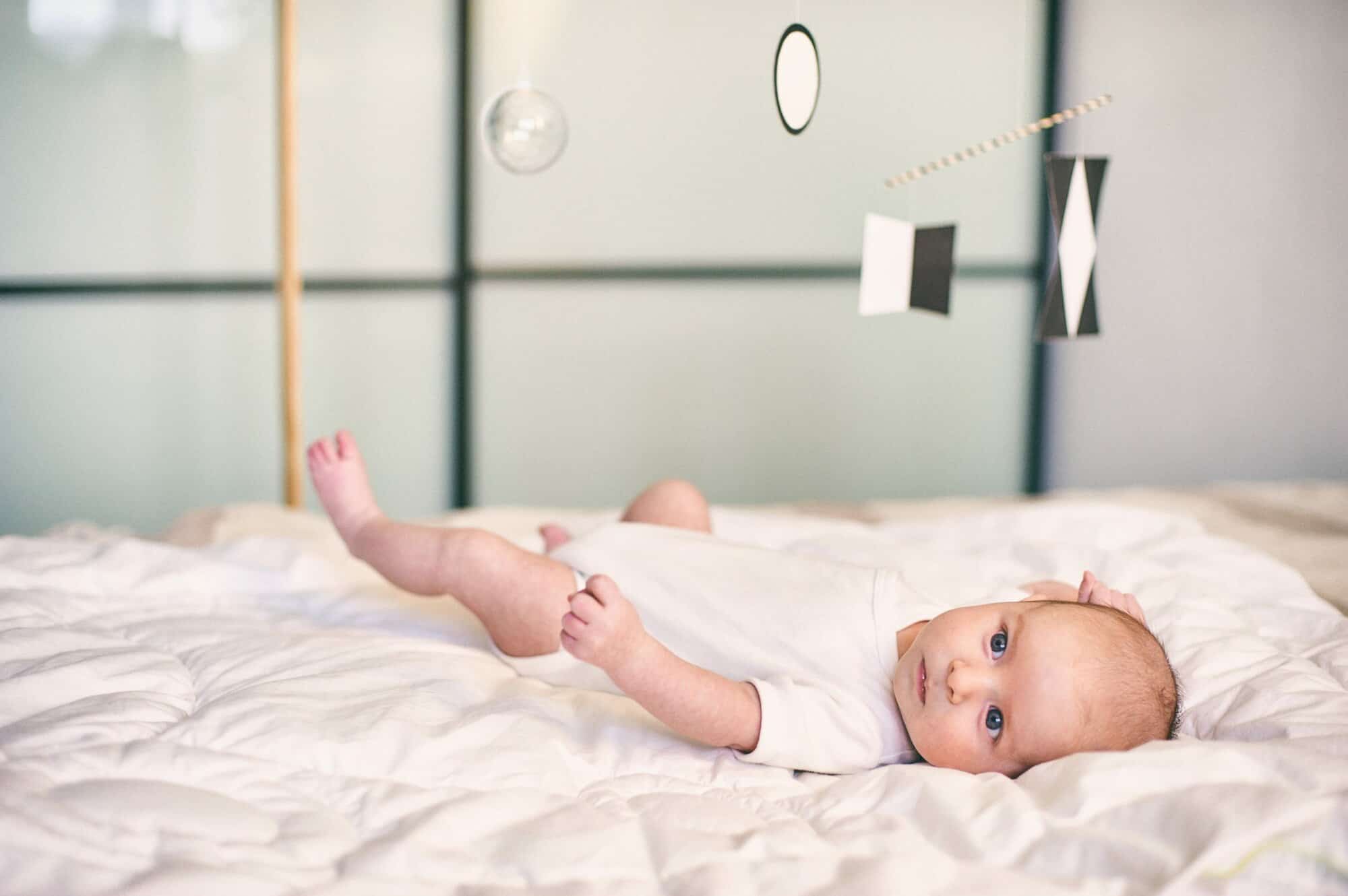 Adorable Baby Boy Infant In White Sunny Bedroom Lying And Looks At Munari Montessori Mobile.