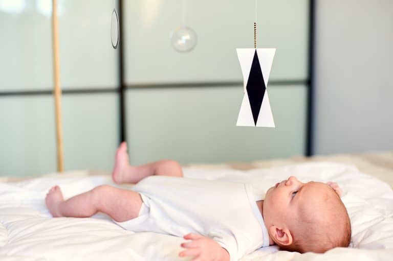 Adorable Baby Boy Infant In White Sunny Bedroom Lying And Looks At Munari Montessori Mobile.