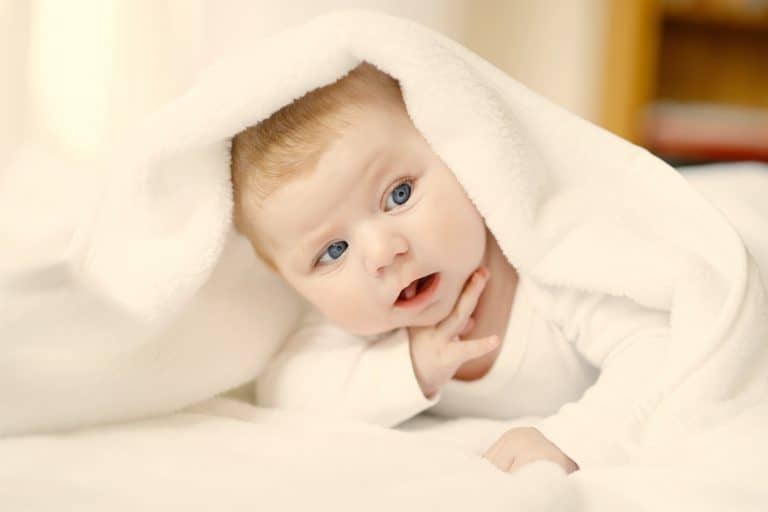 Baby Girl With Blue Eyes Wearing White Towel Or Blanket In White Sunny Bedroom. Newborn Child Relaxi