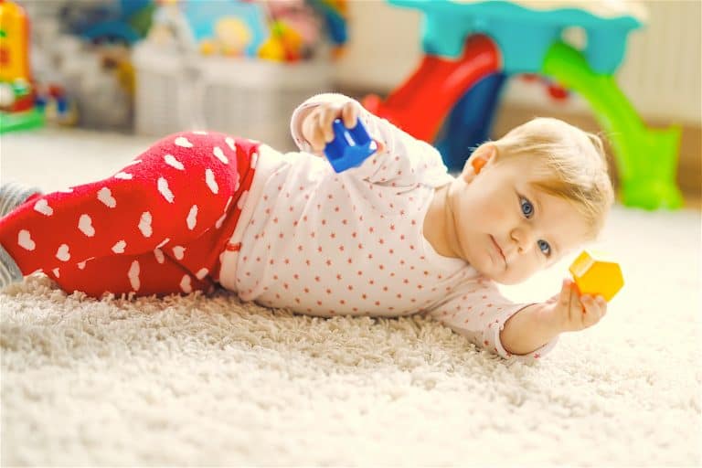 Little Cute Baby Girl Learning To Crawl. Healthy Child Crawling In Kids Room With Colorful Toys. Bac